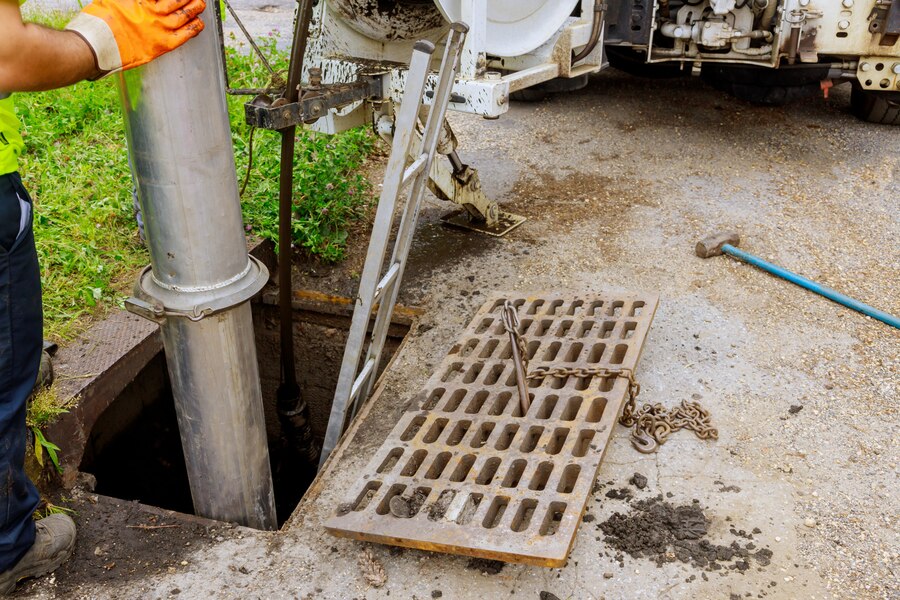 Technicien intervenant sur un réseau d'égout en Belgique : plaque d'égout ouverte avec des canalisations en acier galvanisé, portant des gants de sécurité orange et utilisant du matériel de nettoyage professionnel pour la maintenance du réseau d'assainissement urbain.