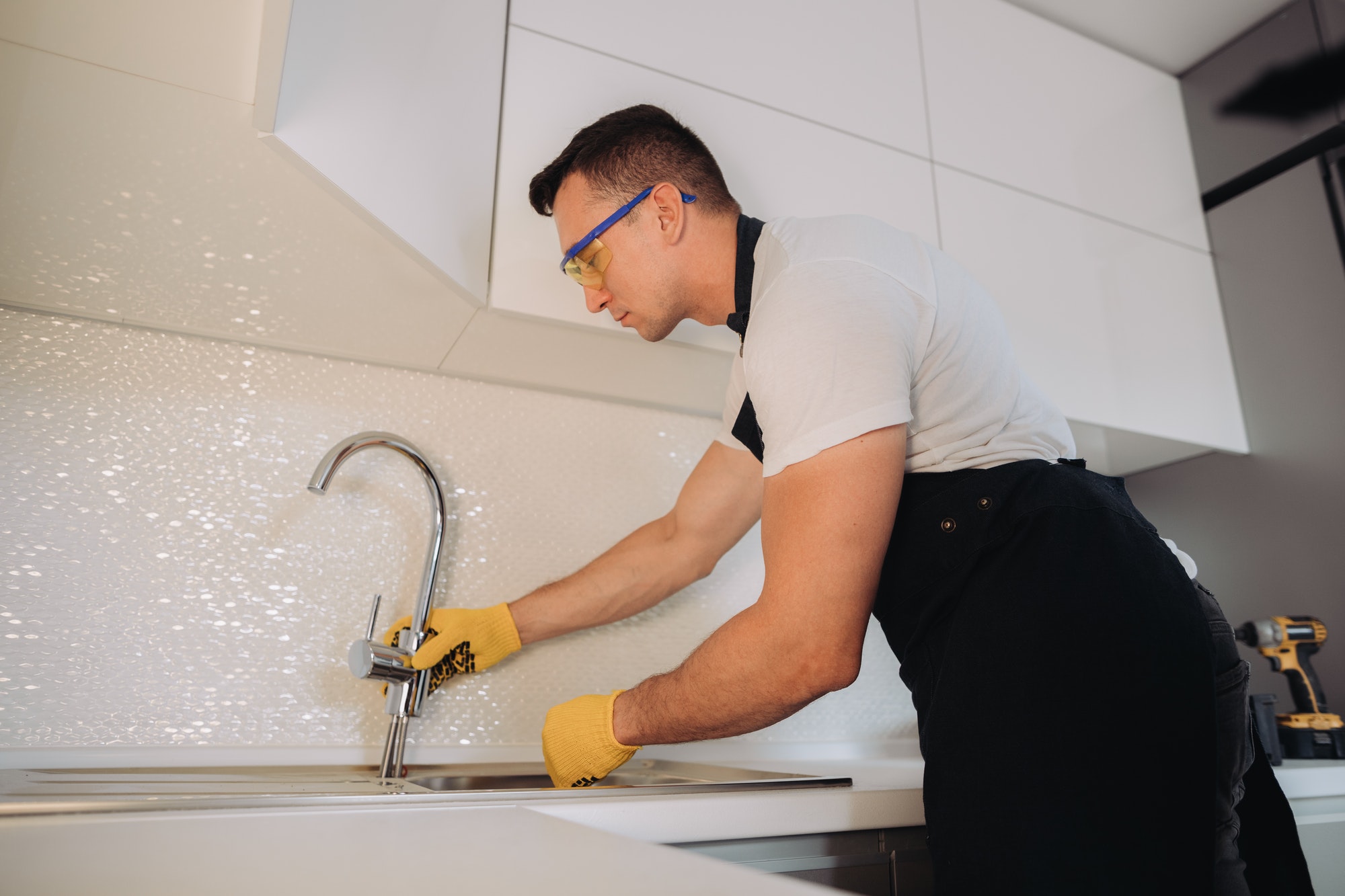 Plombier professionnel en Belgique installant un nouveau robinet dans une cuisine moderne. Il porte des gants de protection jaunes et travaille avec précision pour assurer une installation de plomberie de haute qualité.