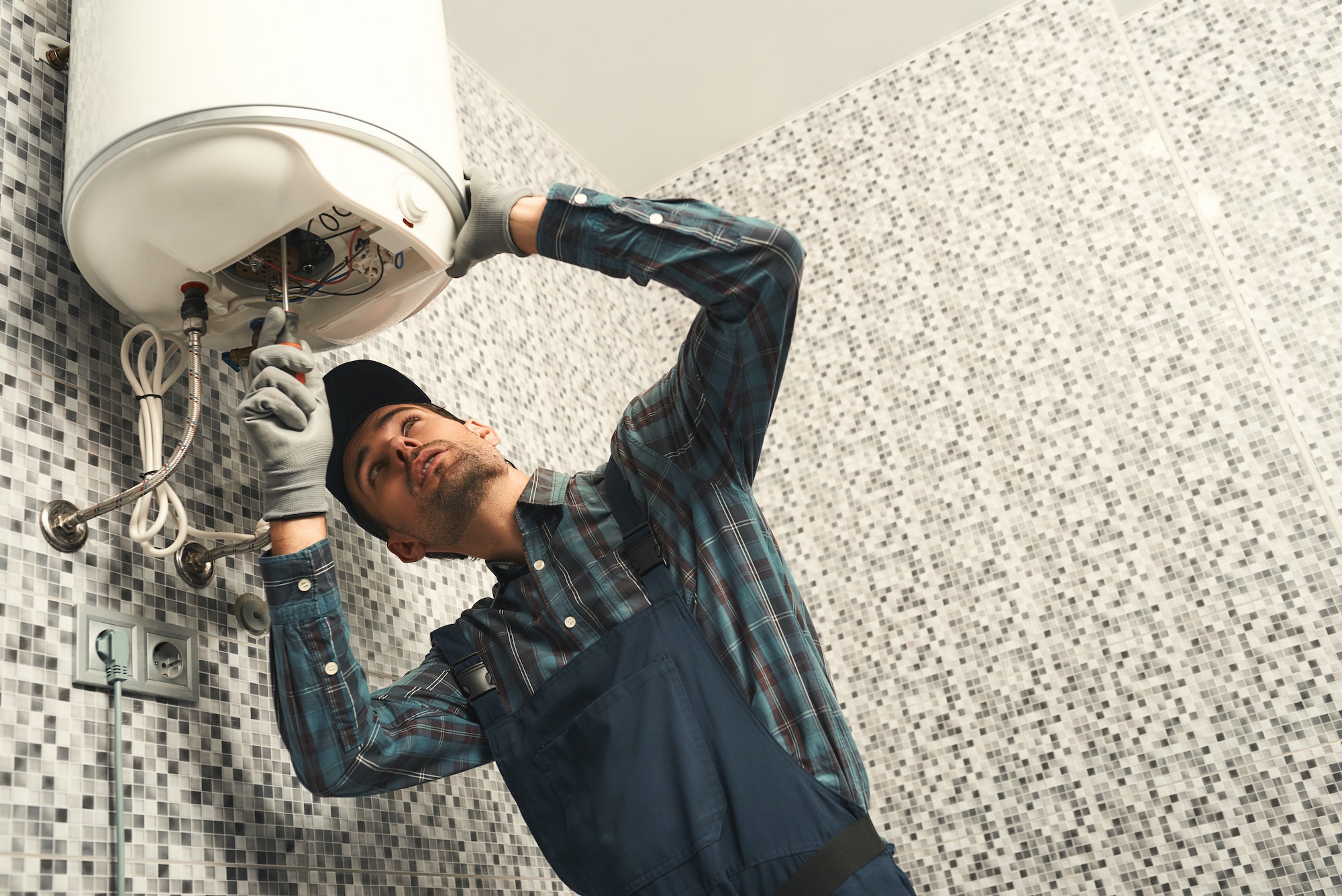 Plombier professionnel en Belgique installant un chauffe-eau électrique mural dans une salle de bain moderne. Il porte un uniforme et des gants de protection tout en vérifiant le système de chauffage pour une installation sécurisée et efficace.