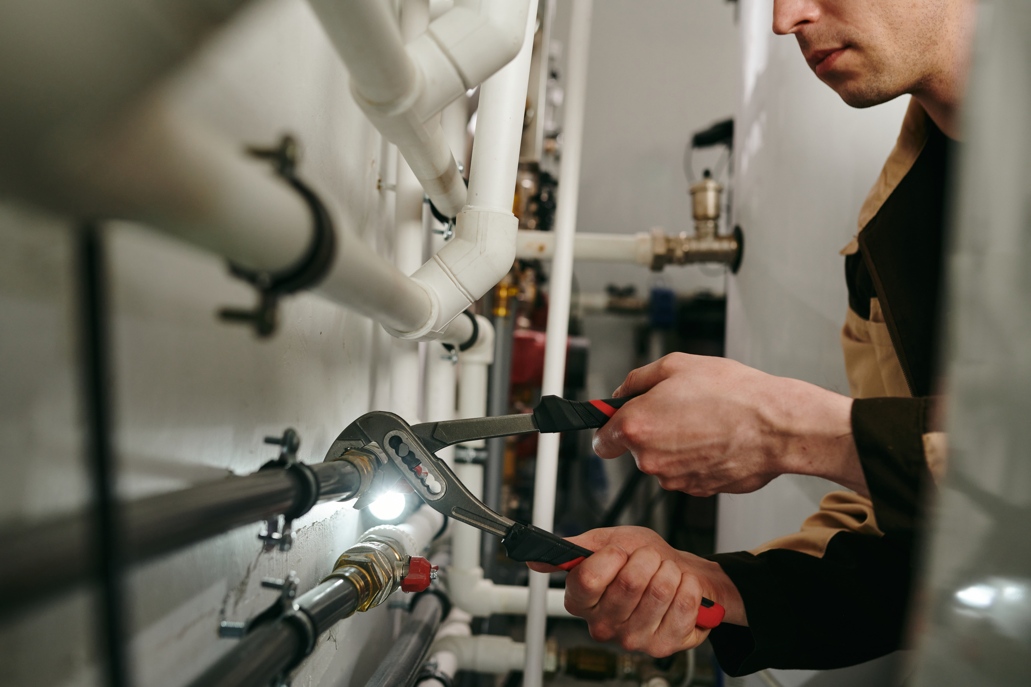 Plombier qualifié utilisant une clé à molette pour réparer une installation de tuyauterie avec des conduites PVC blanches. Vue rapprochée des mains du professionnel effectuant une maintenance sur un système de chauffage dans une chaufferie en Belgique. Éclairage LED professionnel pour un travail de précision.