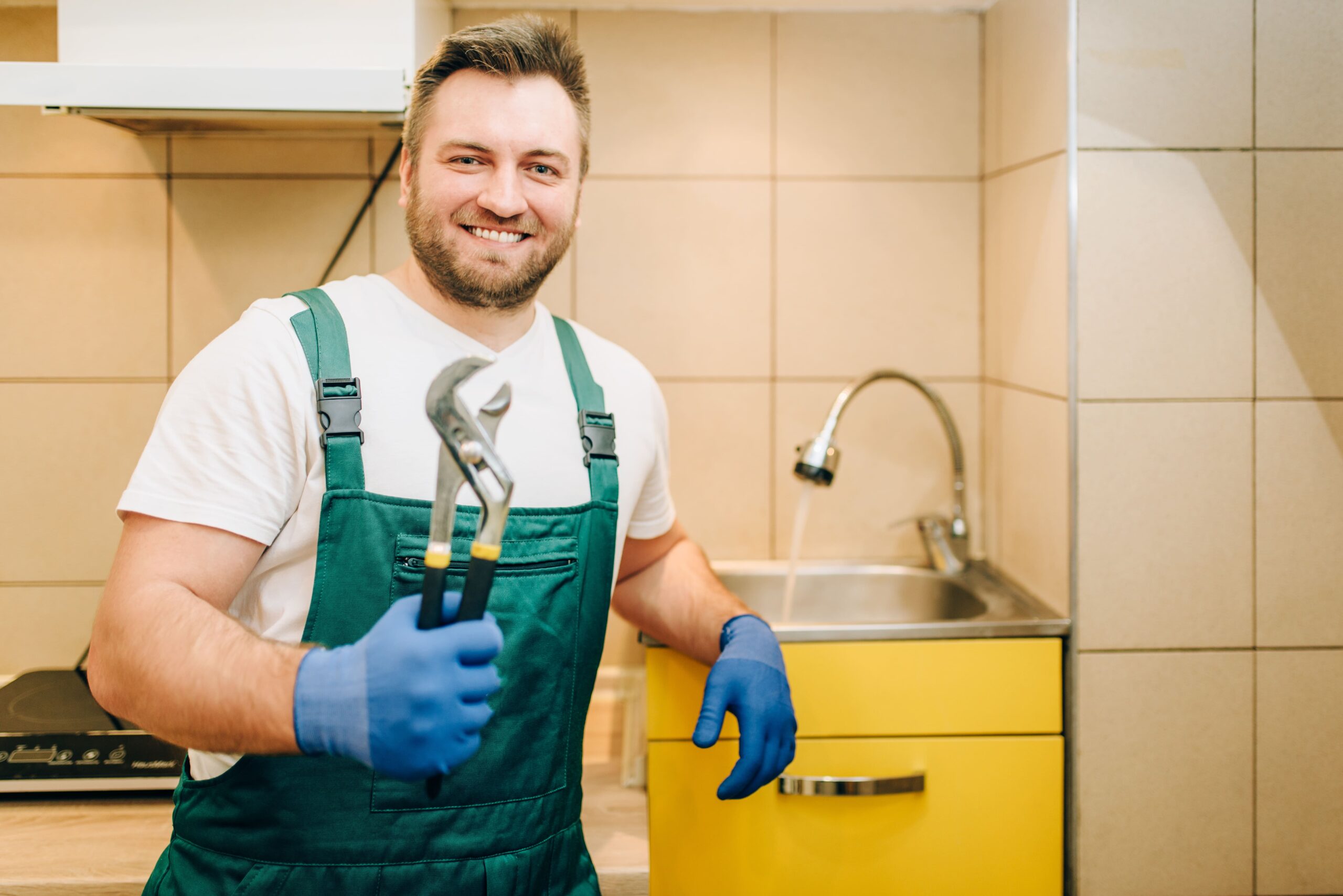 Plombier souriant en uniforme vert tenant une clé à molette devant un évier