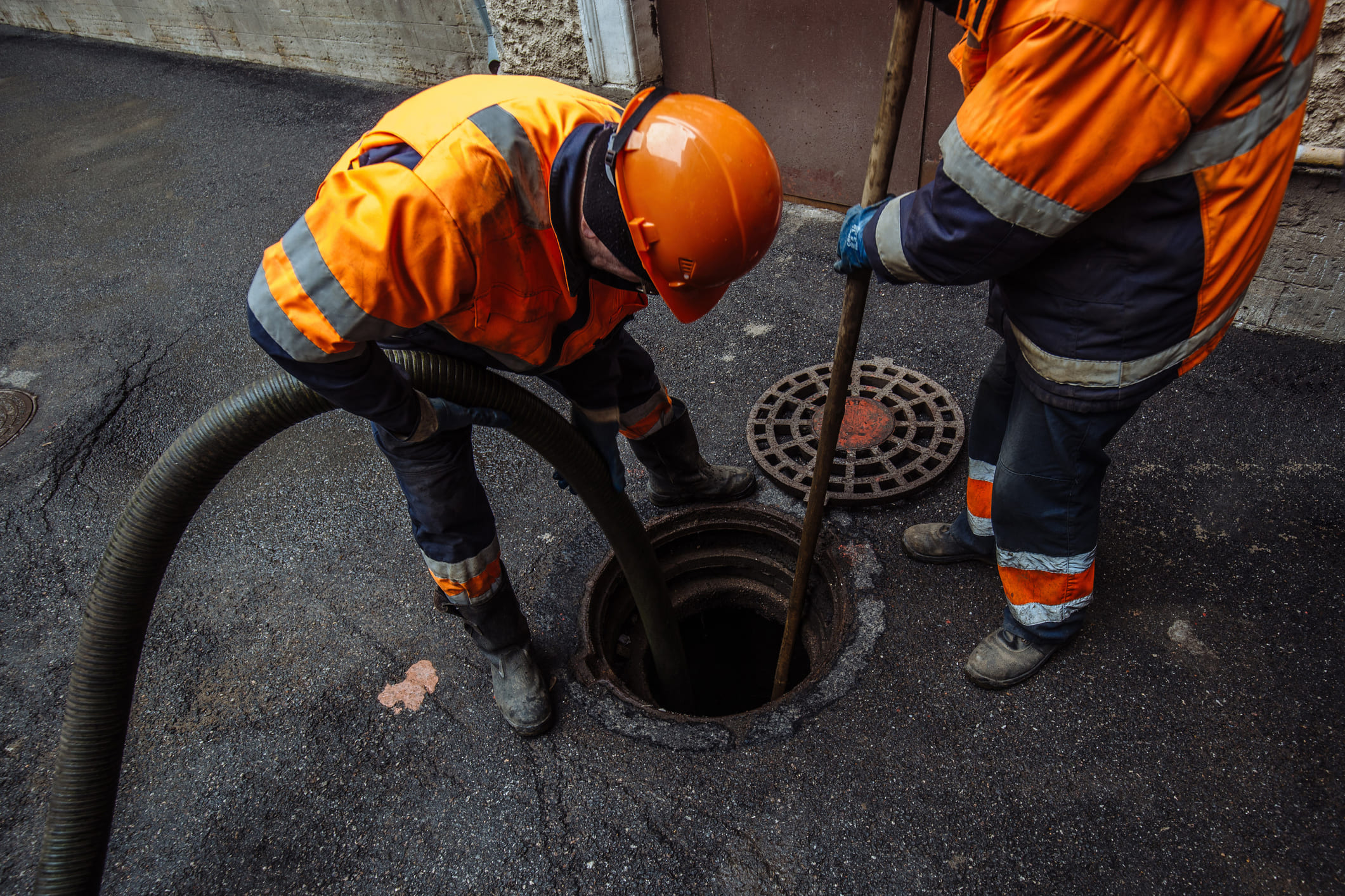 Deux techniciens en tenue orange débouchent une canalisation avec un tuyau et une tige métallique dans une bouche d'égout urbaine.