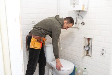 Plombier professionnel en train d'examiner une cuvette WC lors d'un remplacement de toilettes. Il porte une ceinture à outils et inspecte soigneusement l'installation dans une salle de bain carrelée de blanc.